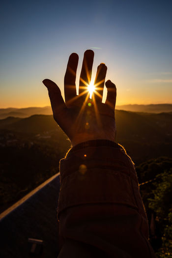 Cropped hand of person against sun during sunset