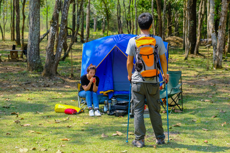 Rear view of man with hiking pole standing near friend sitting by tent in forest