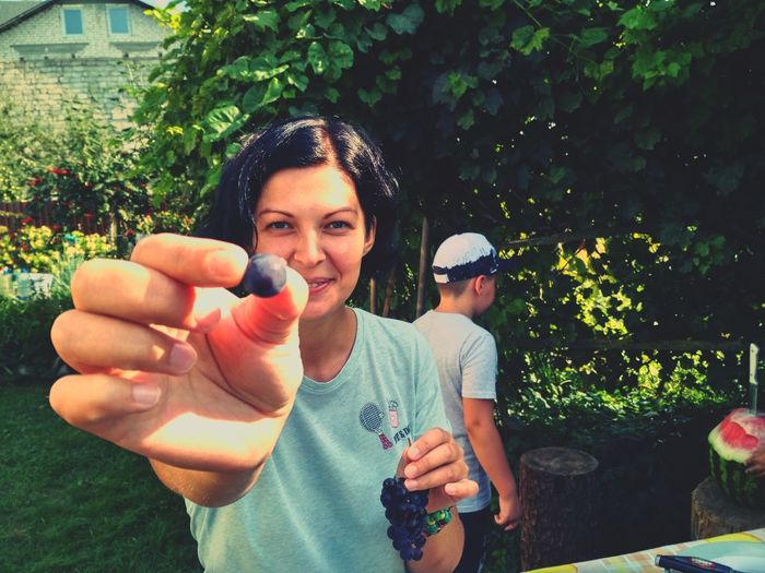 Portrait of smiling young woman holding grape while standing with son in yard