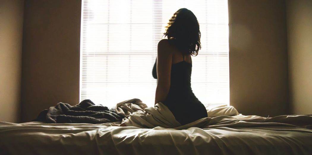 Rear view of young woman looking through window while sitting on bed