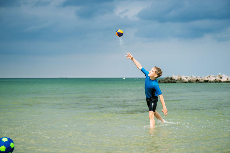 Full length of boy playing rugby ball and soccer ball in water at beach