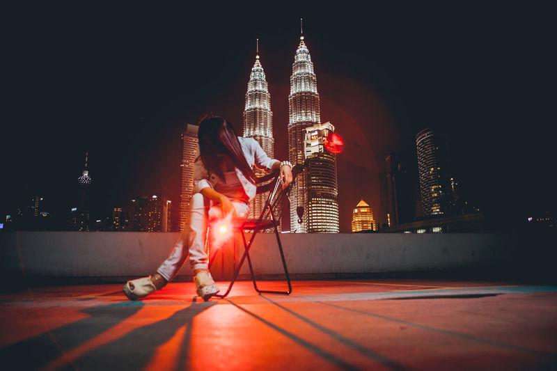 Low angle view of woman sitting on chair against illuminated building in city at night
