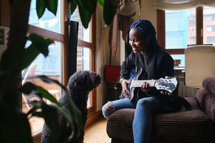 Smiling african american young female musician with dreadlocks wearing jacket and jeans playing guitar while sitting on sofa opposite poodle dog in living room
