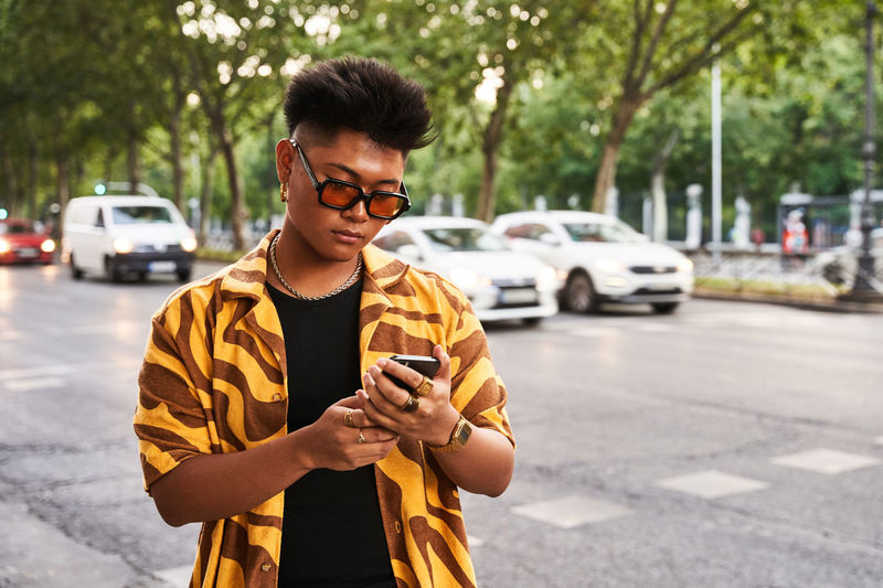 Modern asian male in trendy orange shirt and sunglasses checking smartphone while standing on street