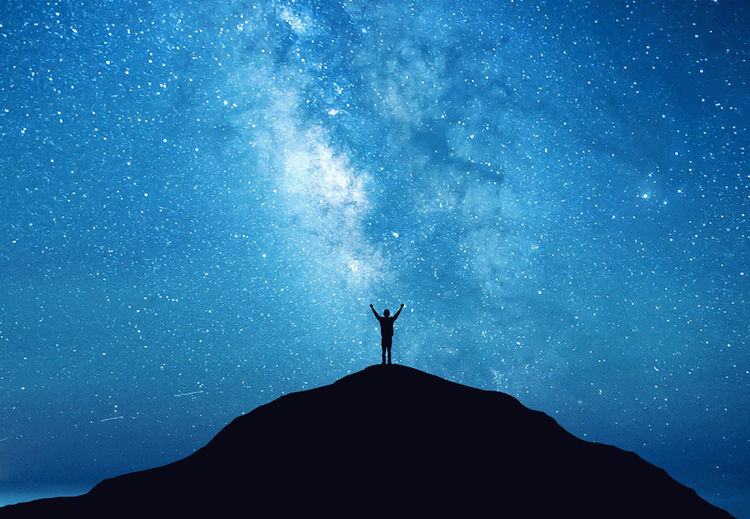 Low angle view of silhouette man standing on mountain against blue star field at night