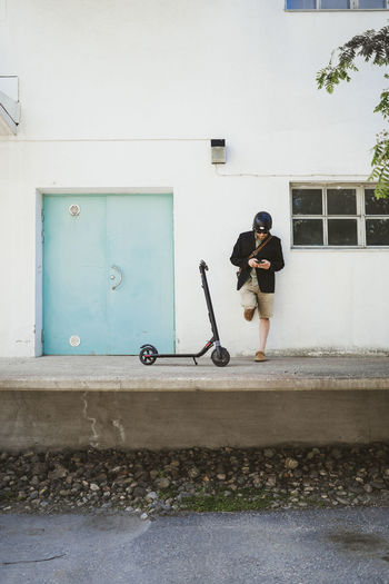 Man with electric scooter leaning against facade using smartphone