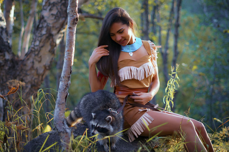 Smiling young woman in traditional clothing sitting by raccoon in forest
