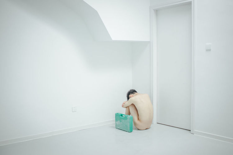 Naked man with suitcase sitting on floor against wall
