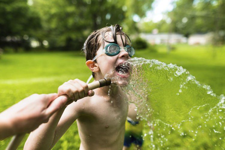 Shirtless boy wearing swimming goggles while playing with water at yard
