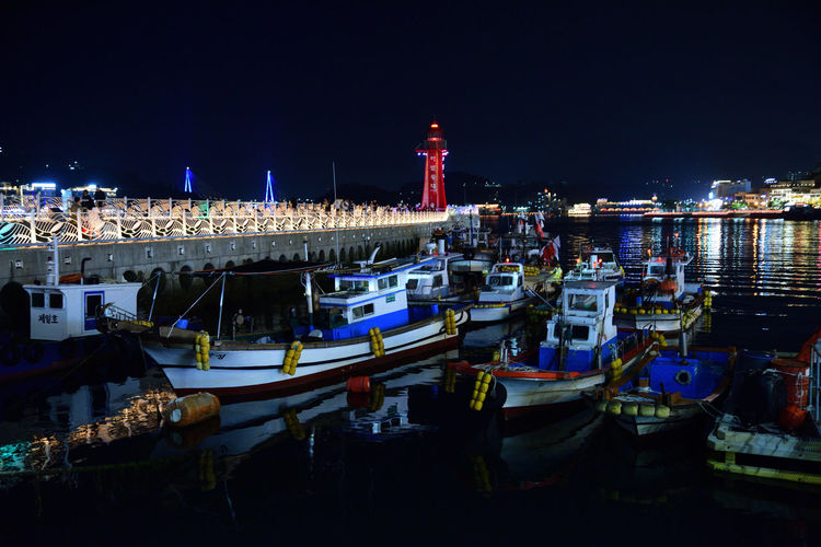 Boats moored at harbor against illuminated buildings in city at night