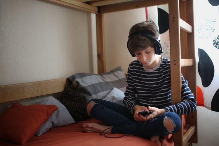 Teenager with a phone and headphones listening to music on the first floor of a bunk bed, 