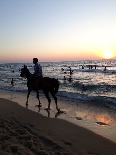 People riding horse on beach against sky during sunset
