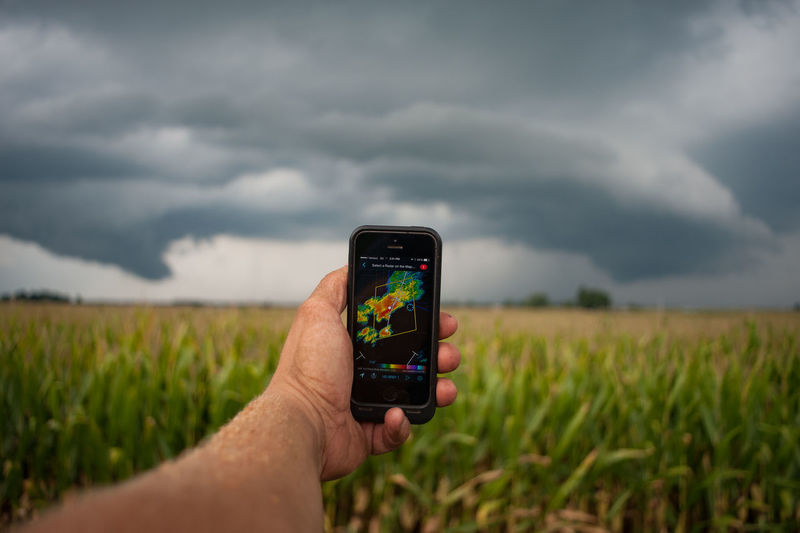 Cropped hand holding smart phone in field against cloudy sky