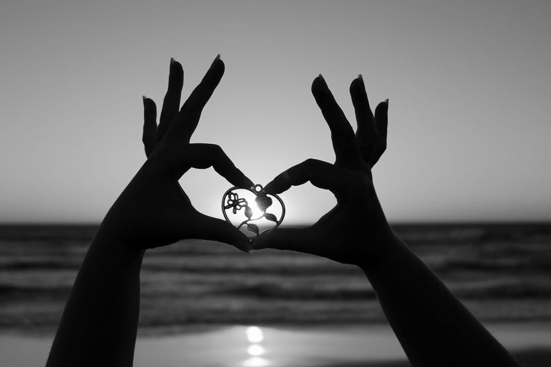 Cropped image of silhouette hands holding heart shaped locket against sky