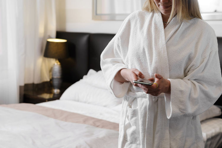 Woman wearing bathrobe while standing in hotel room using smartphone. communicating, 