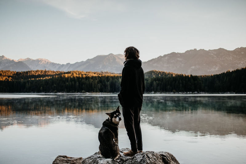 Girl is standing on a rock with her dog at lake Eibsee Berchtesgaden , Germany Eibsee Adventuers Adventure Alps Animal Beauty In Nature Dog Lake Leisure Activity Lifestyles Mammal Mountain Mountain Range Mountains One Person Outdoors Pet Owner Real People Rear View Standing Water