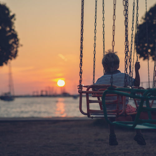 Rear view of boy sitting on swing at beach against sky during sunset