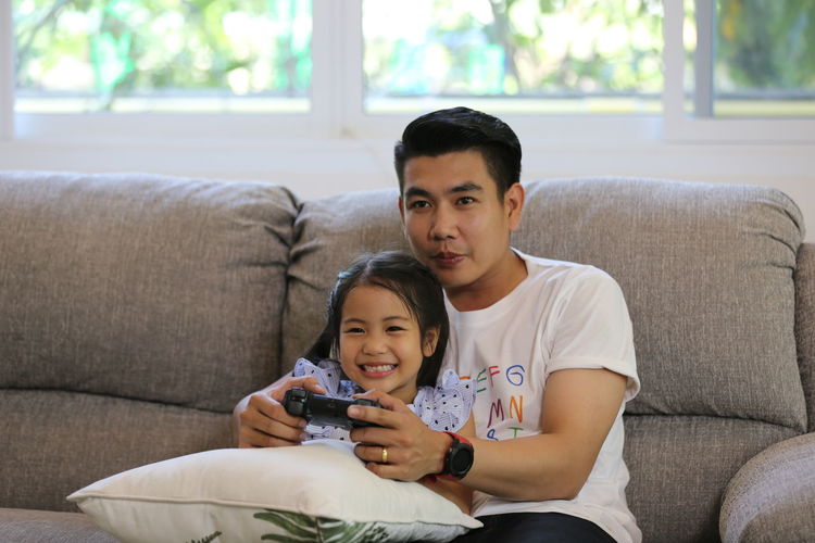 Father and daughter playing video game while holding remote control on sofa
