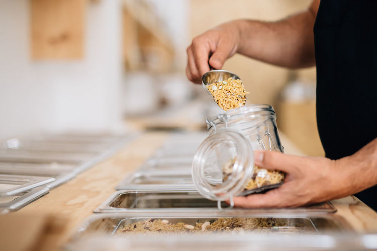 Side view of anonymous female pouring dry pasta inside glass jar in local store