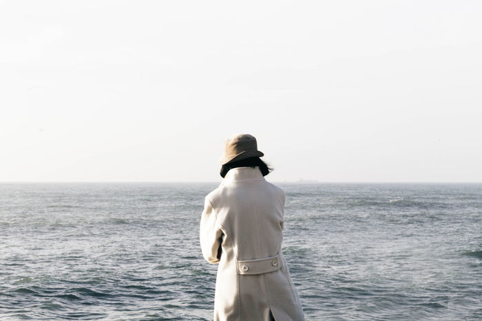 Rear view of person in long coat standing by sea against sky