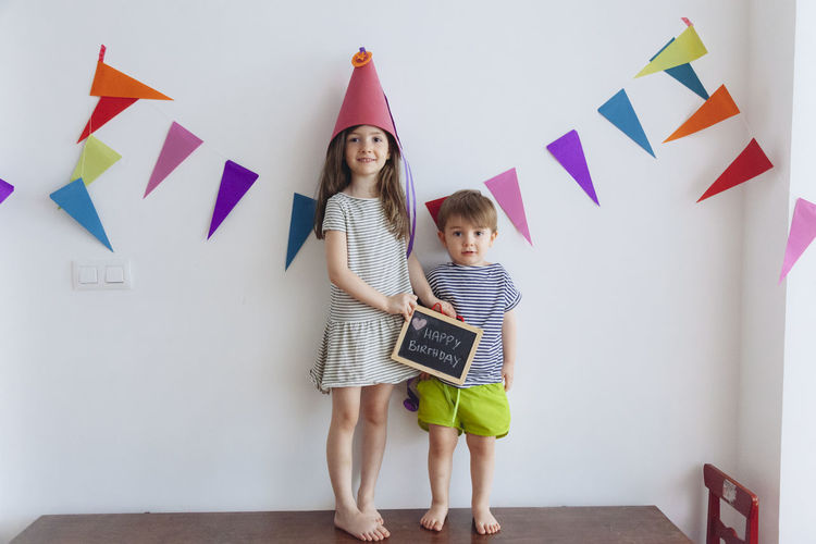 Portrait of cute sister wearing party hat standing with boy on table