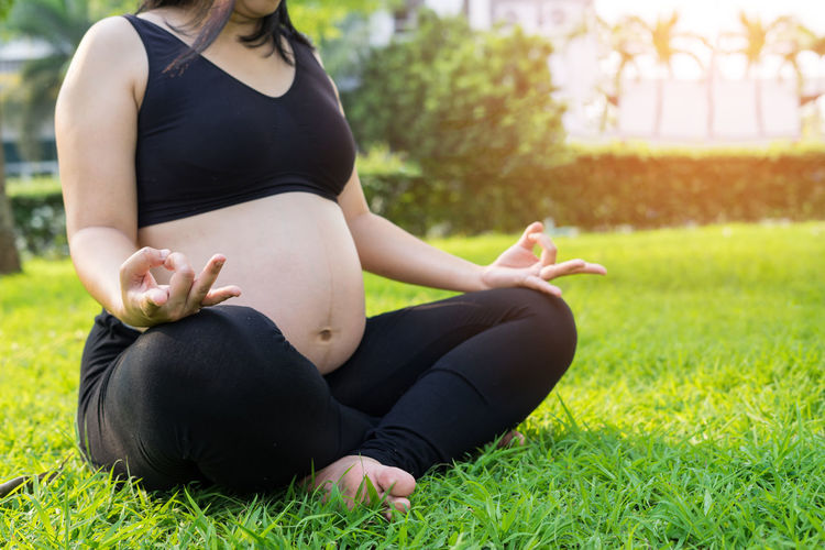 Low section of pregnant woman meditating while sitting on grassy field at park