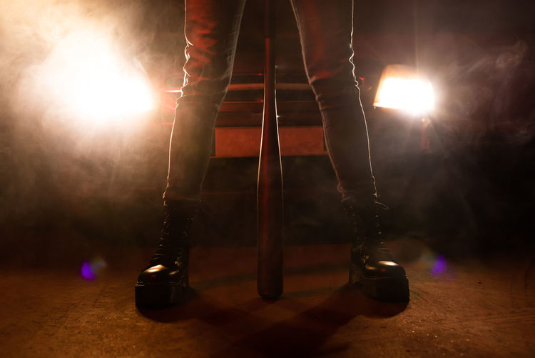 Crop legs of faceless woman in jeans and black boots with massive sole holding bat with car headlights on background