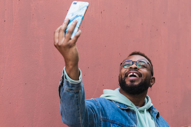 Low angle cheerful adult black hipster guy in casual outfit taking self portrait on mobile phone against red background