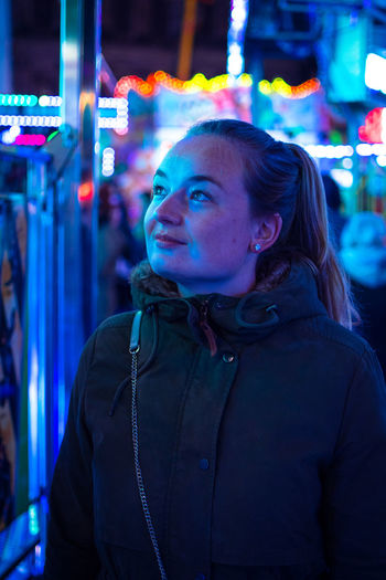 Close-up of young woman standing against illuminated amusement ride at night