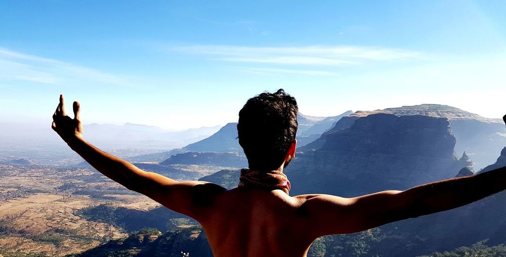 Rear view of shirtless man with arms outstretched against sky