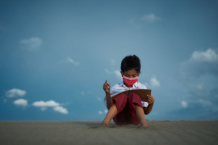 Little boy sitting and reading book at the beach under blue sky