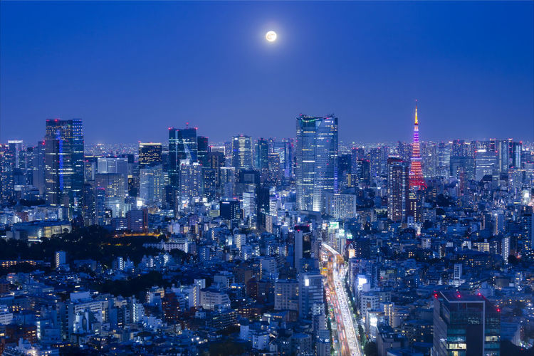 Illuminated cityscape against the full moon and clear blue sky