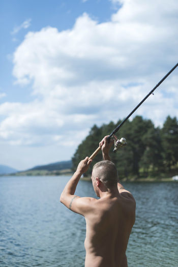 Shirtless man fishing in lake against sky