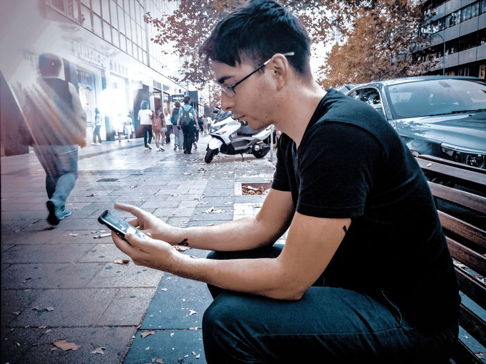 Side view of man using phone while sitting on bench at sidewalk in city