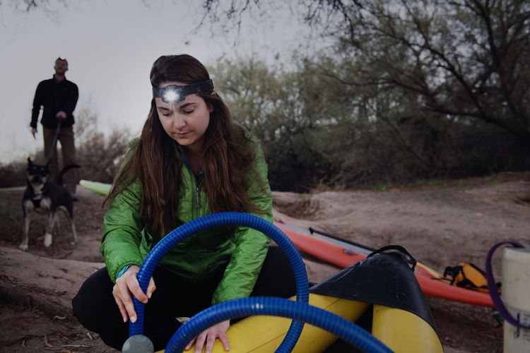 Woman inflating kayak with friends holding dog leash in background at forest
