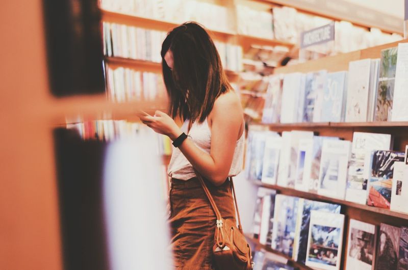 Side view of woman using mobile phone while standing amidst bookshelves at store