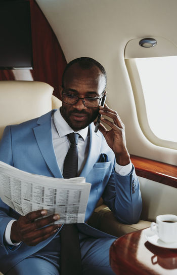 Young businessman reading newspaper while talking on smart phone in airplane