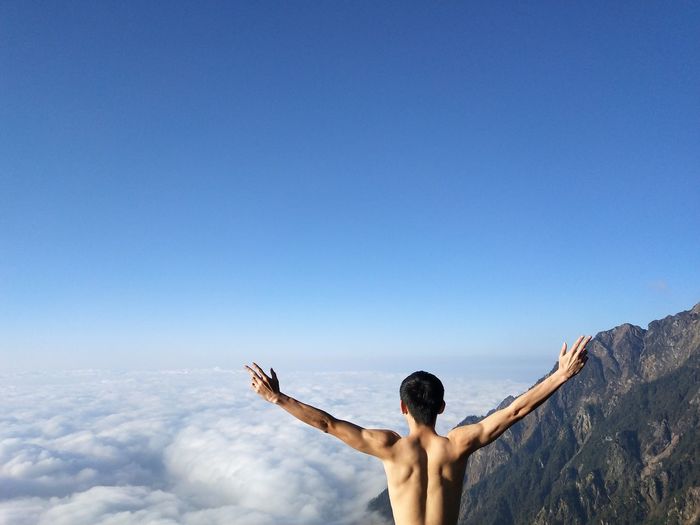 Rear view of shirtless man with arms outstretched standing against blue sky