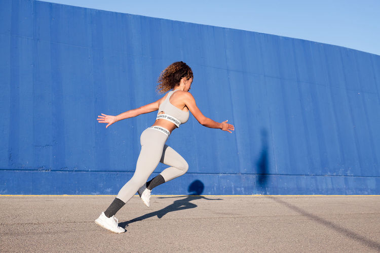 Side view of focused female runner in sports clothes sprinting on pavement with shades against blue wall during workout