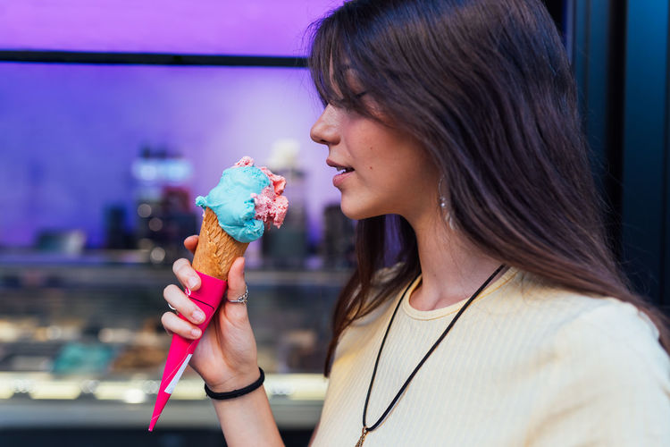 Crop cheerful young female in pendant and earrings with delicious gelato in waffle cone looking away on street