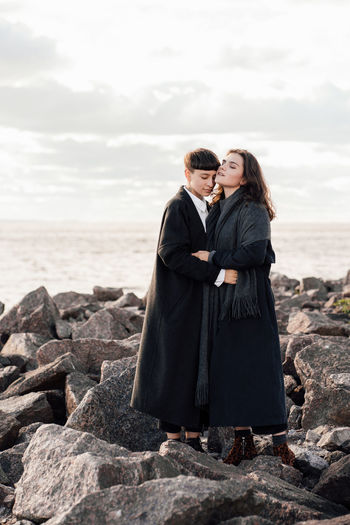 Lesbian women embracing while standing on rock against sea and sky