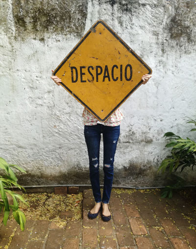 Low section of woman holding road sign while standing against wall