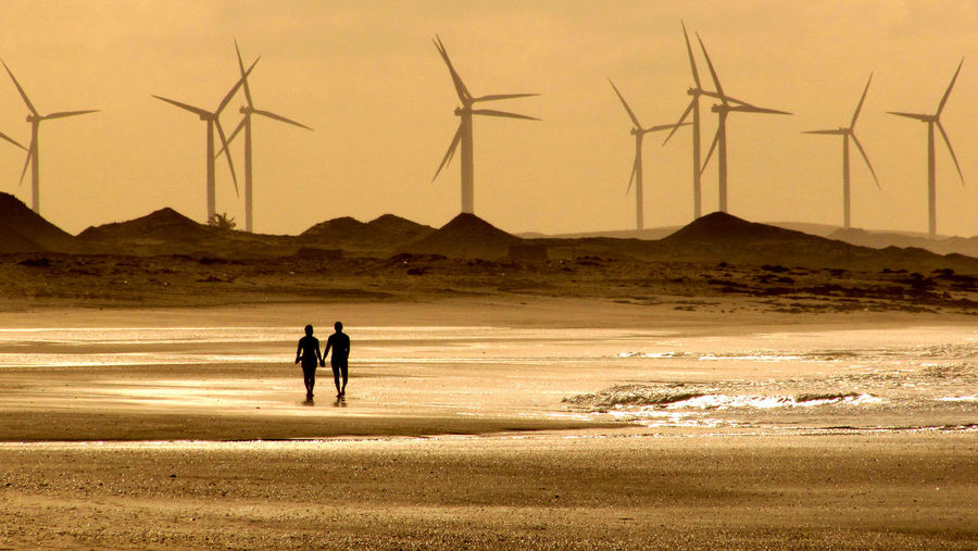 Silhouette couple walking on beach against windmills during sunset