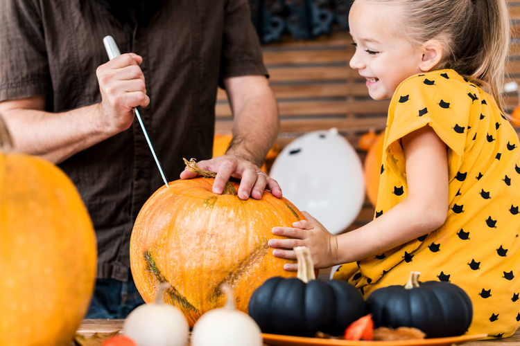 Midsection of father cutting pumpkin with girl sitting a home