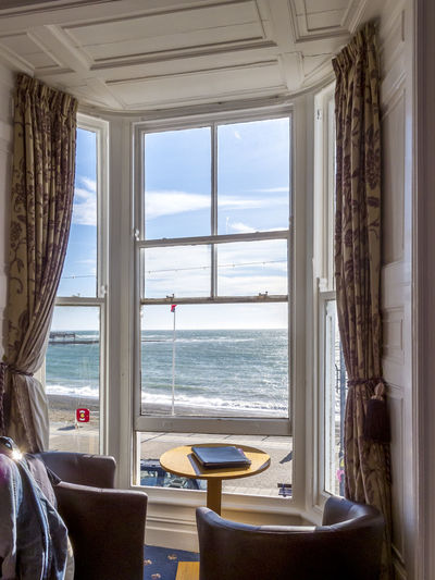 View on the sunlit sea through a window framed by curtains, table and armchairs in the foreground 