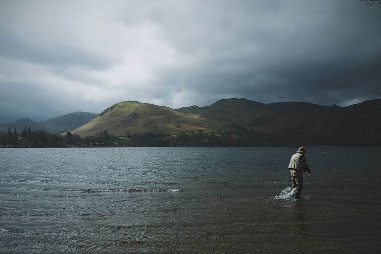 Man walking in river against mountains and cloudy sky at dusk