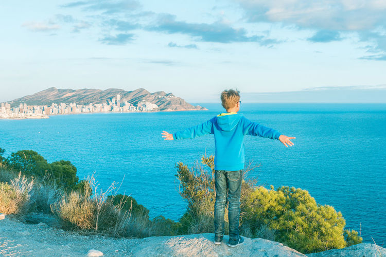 Rear view of boy standing with arms outstretched while looking at sea against sky