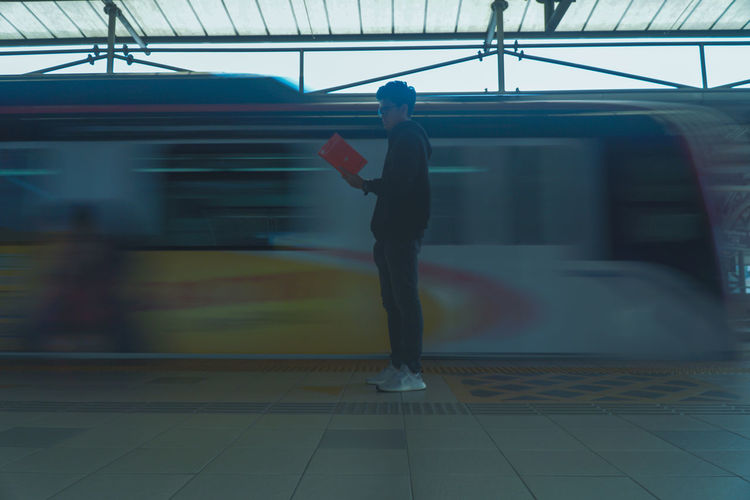 Side view of man reading book while standing against train moving at railroad station
