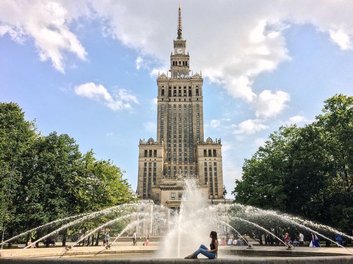 Side view of woman sitting by fountain in front of palace of culture and science