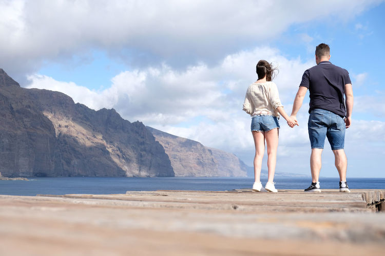 Rear view of couple holding hands while standing at beach against sky